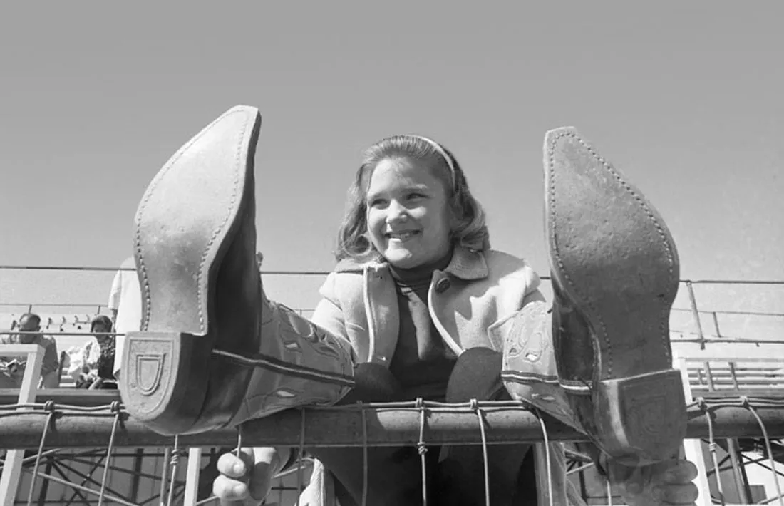 A child with cowboy boots propped up on a fence, smiling
