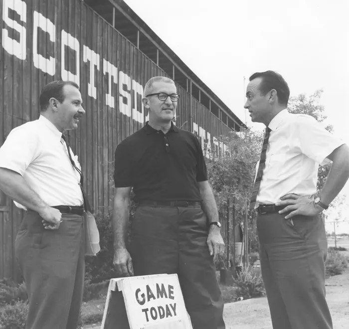 Three men standing together in front of a building reading "Scottsdale Stadium" and a sign that reads "Game Today"